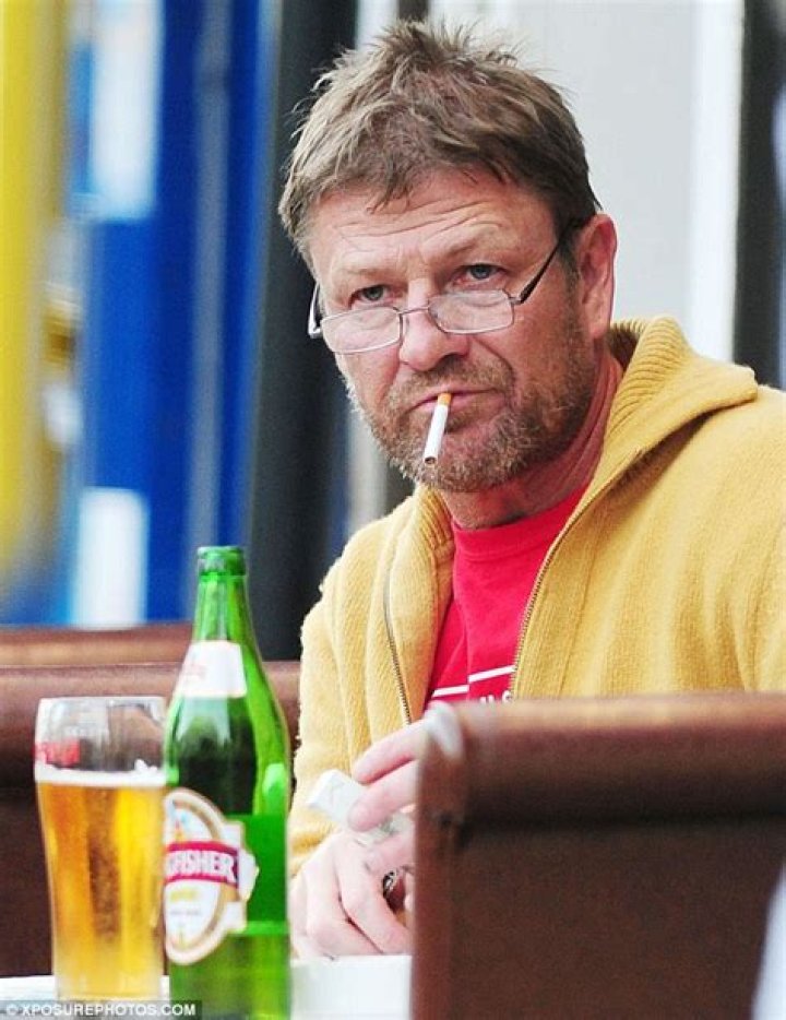 Sean Bean looks disheveled as he sits alone drinking a bottle of lager and smoking cigarettes